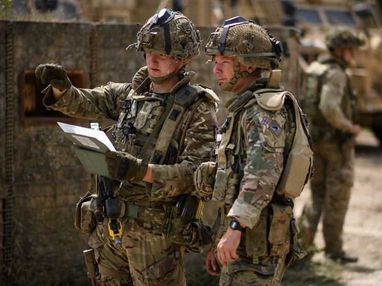 Two British soldiers in camouflage uniforms and helmets examining documents with military vehicles in background