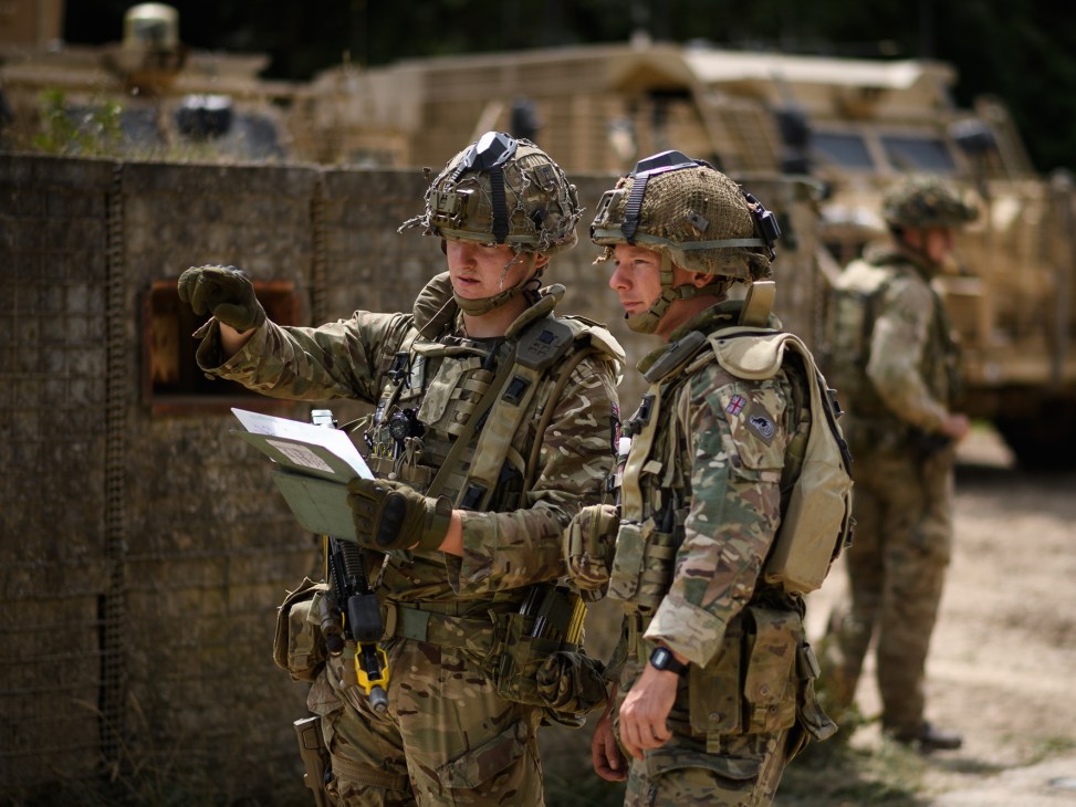 Two British soldiers in camouflage uniforms and helmets examining documents with military vehicles in background