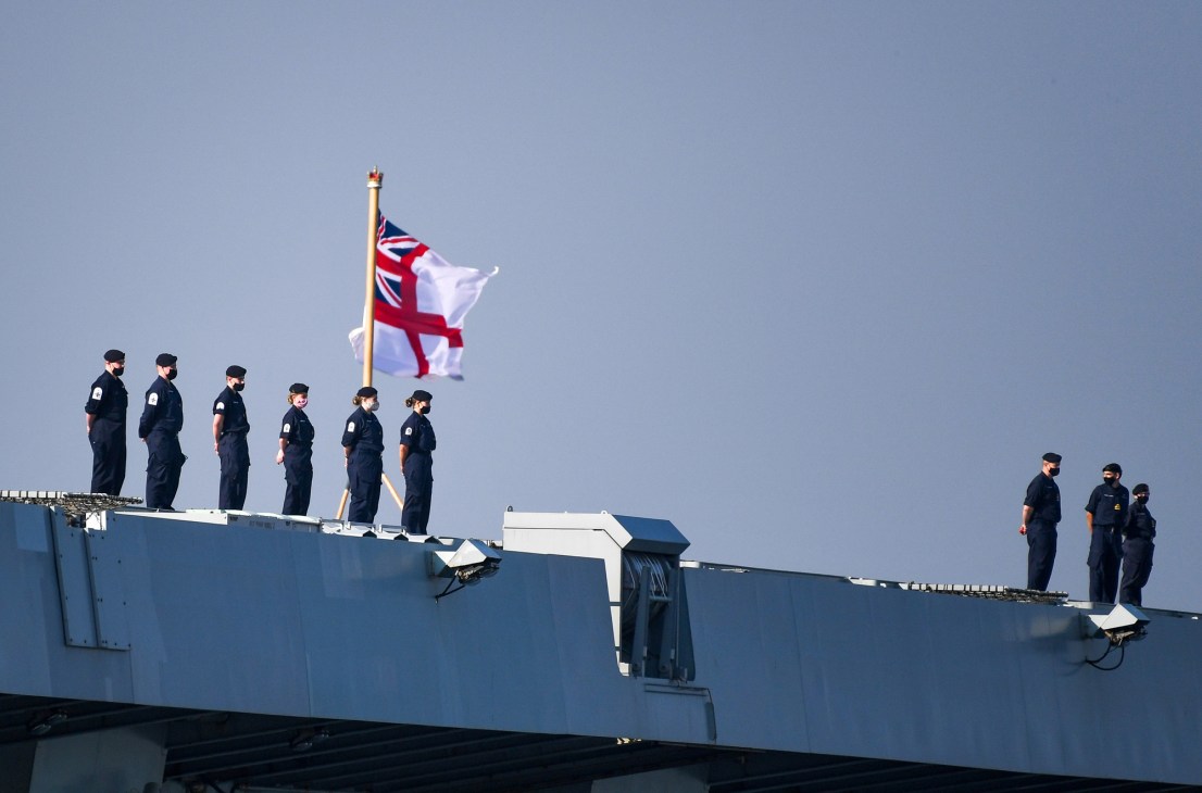 Naval personnel in dark uniforms standing on ship deck with British naval ensign flag flying