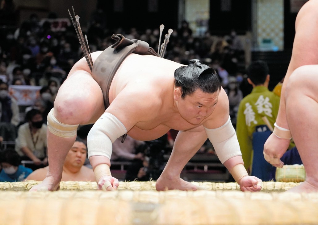 A sumo wrestler in traditional white loincloth and topknot performs in the ring before a crowd of spectators.