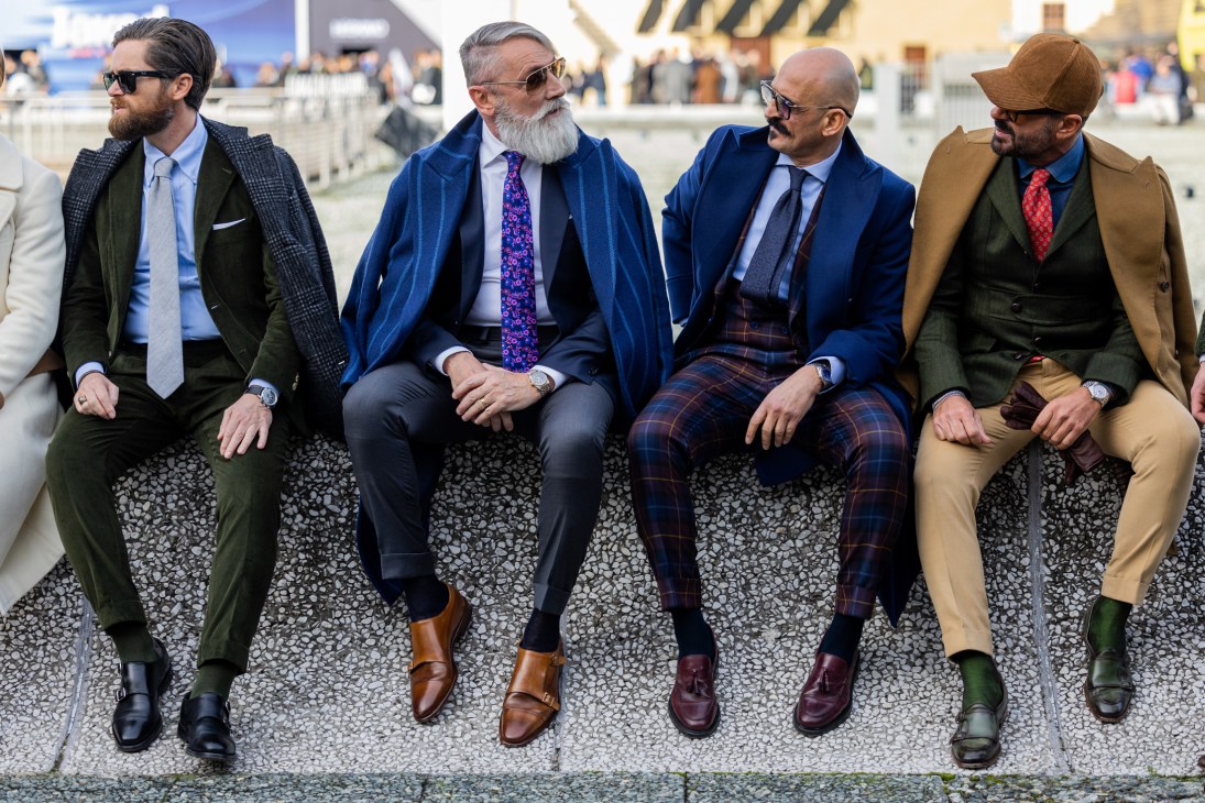 Four well-dressed men in tailored suits and loafers sitting on a ledge at what appears to be a fashion event