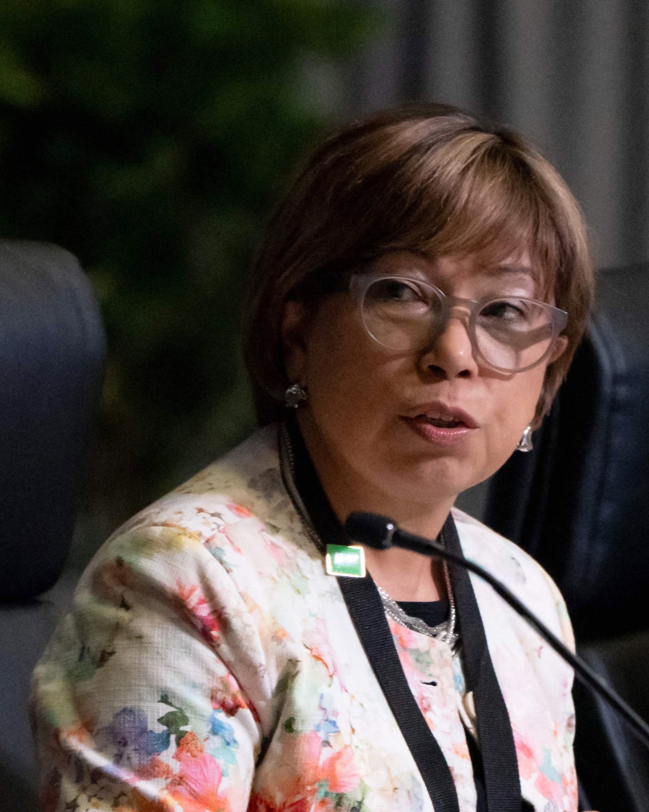 Woman with glasses and floral blazer speaking at podium with microphone in formal setting
