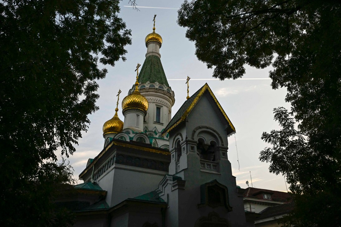 Russian Orthodox church with golden domes and green roof surrounded by trees.
