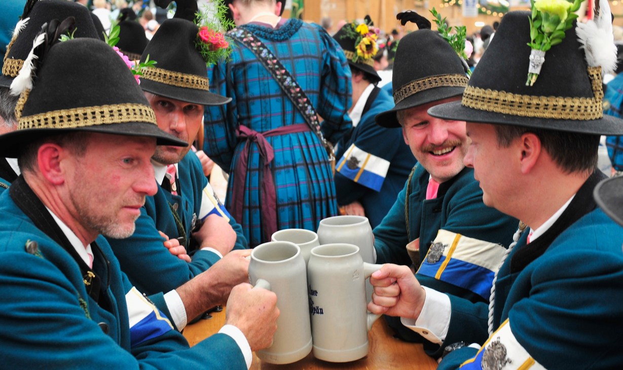 Men in traditional Bavarian attire with black hats and gold trim toasting with beer steins.