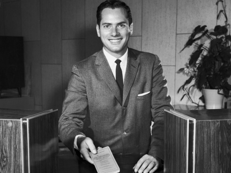 Portrait of a man in a grey suit and dark tie holding a document at a desk in an office setting.