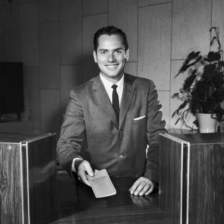 Portrait of a man in a grey suit and dark tie holding a document at a desk in an office setting.