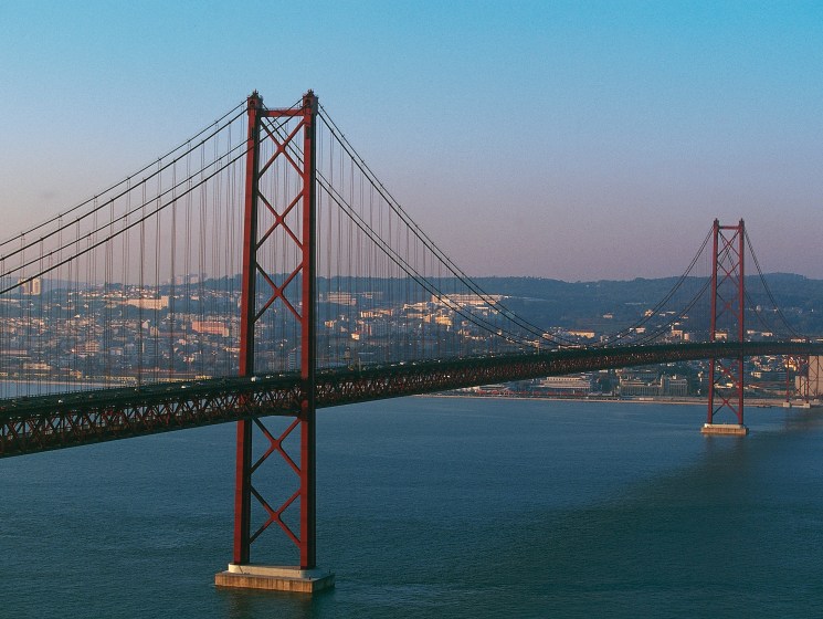 Red suspension bridge spanning blue water with hillside cityscape in background