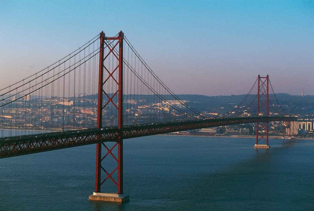 Red suspension bridge spanning blue water with hillside cityscape in background