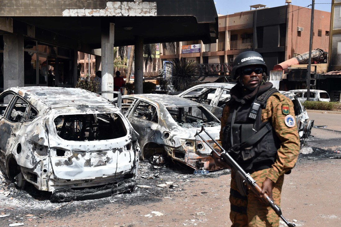 Armed gendarme stands guard near burnt-out vehicles in Burkina Faso urban street.