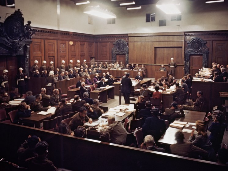 Black and white photo of the Nuremberg trial courtroom showing defendants' dock, judges' bench and packed gallery