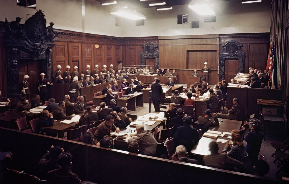 Black and white photo of the Nuremberg trial courtroom showing defendants' dock, judges' bench and packed gallery