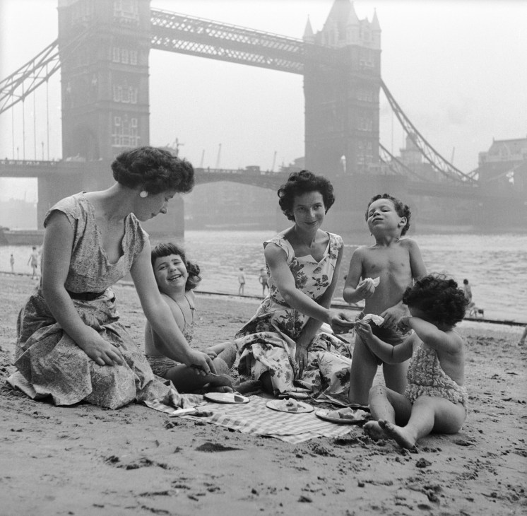 Two women and three children enjoying a picnic on a sandy beach with Tower Bridge visible in the background