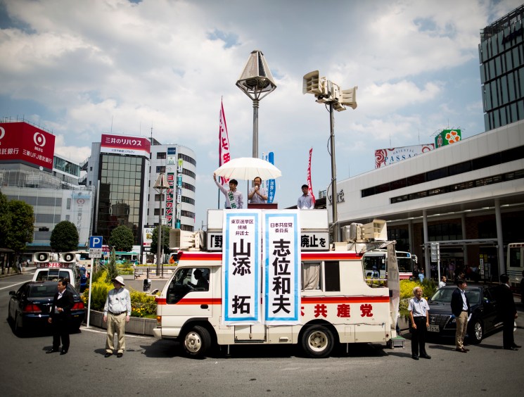 Japanese Communist Party politicians campaign from the roof of a van outside a busy urban train station.