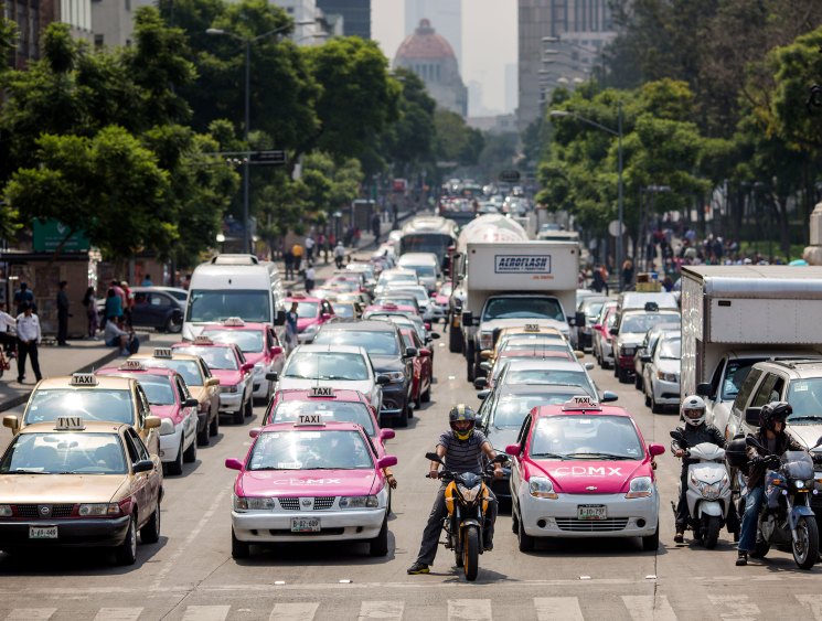 Heavy traffic congestion with numerous taxis, cars, and motorcycles gridlocked on a tree-lined urban street