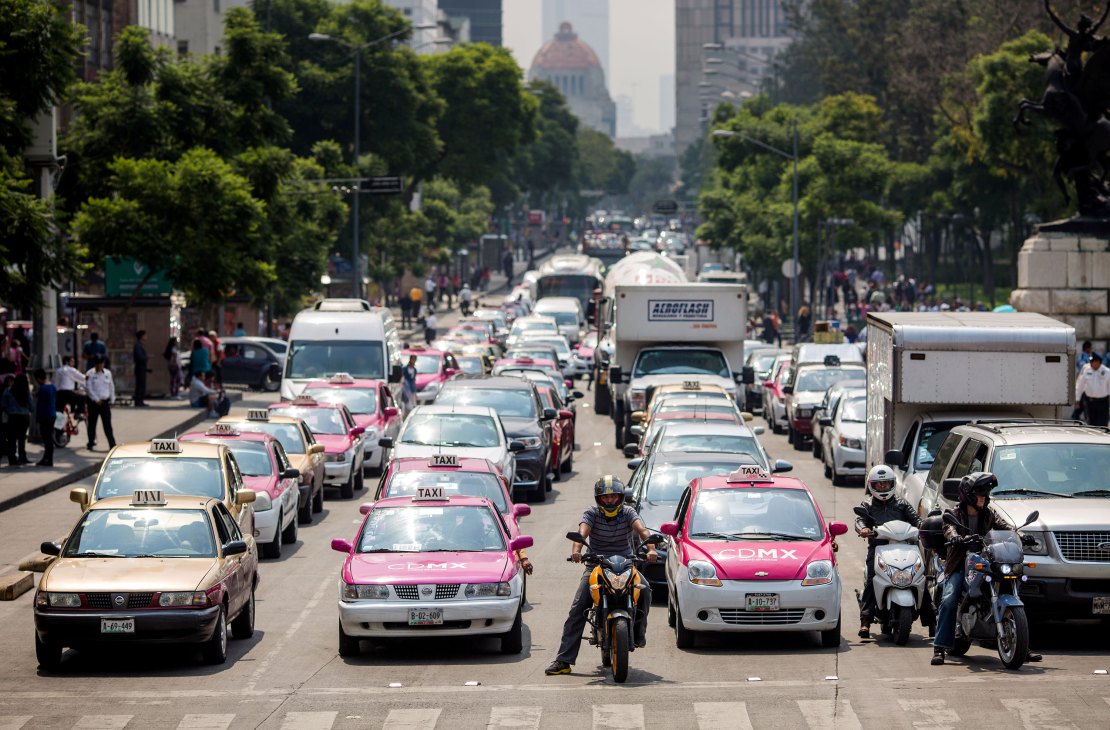 Heavy traffic congestion with numerous taxis, cars, and motorcycles gridlocked on a tree-lined urban street
