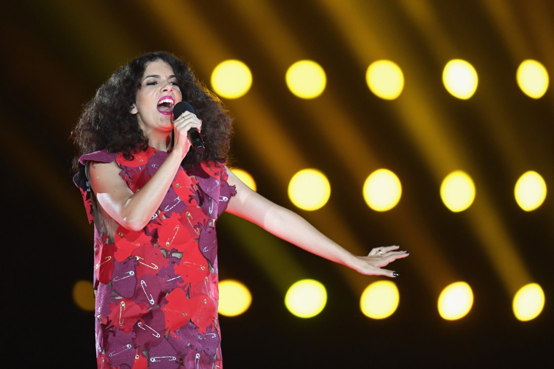 Singer performing on stage with microphone, wearing red patterned outfit against backdrop of bright stage lights