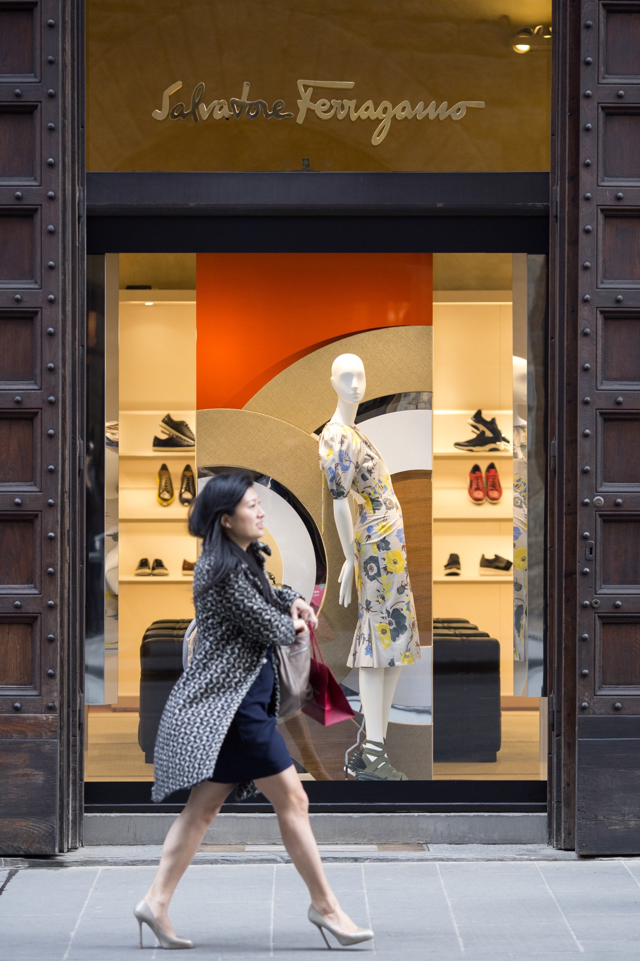 Woman in patterned coat walking past Salvatore Ferragamo storefront with window display