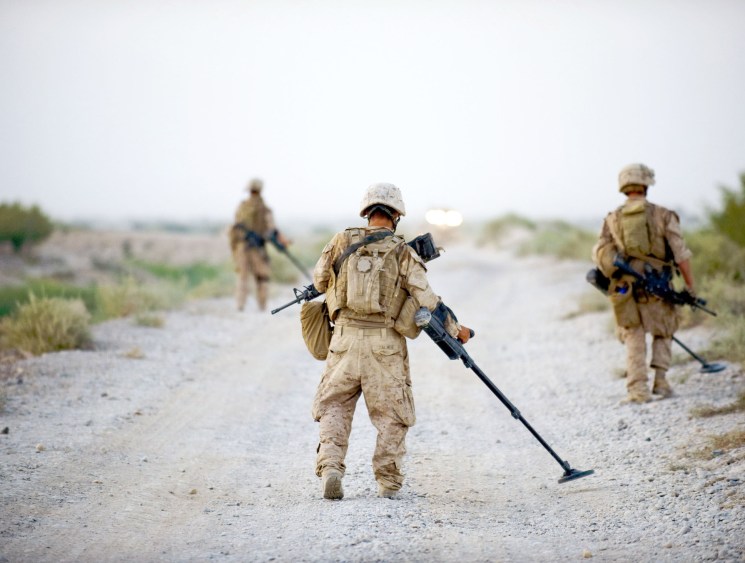 Three soldiers in desert camouflage patrol a dusty road, one carrying a metal detector to sweep for improvised explosive devices.