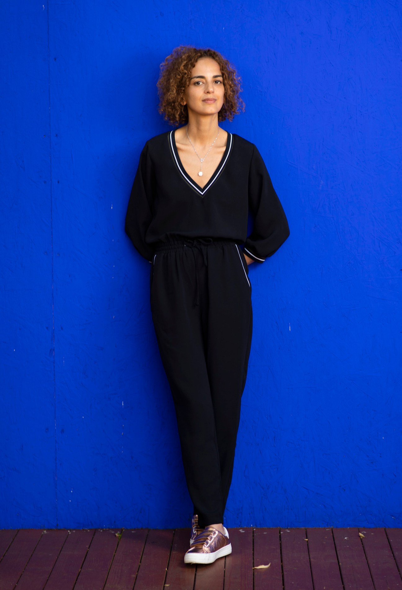 Leïla Slimani, with curly reddish-brown hair wearing a black v-neck sweater with white trim and black pants stands against a bright blue wall, looking off to the side.