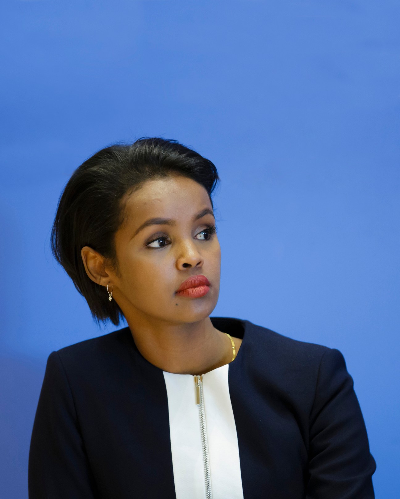 Portrait of a woman with short dark hair wearing a navy blazer with white shirt against a blue background.