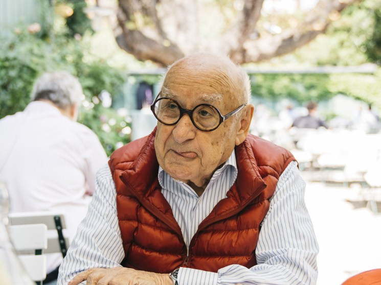 An elderly man in round glasses and a red puffer vest sits at an outdoor café table with an orange cap beside him.