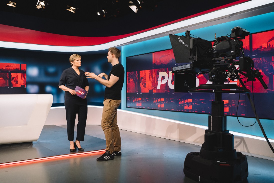 TV presenter Corinna Milborn being interviewed on a modern studio set with red and blue lighting design.