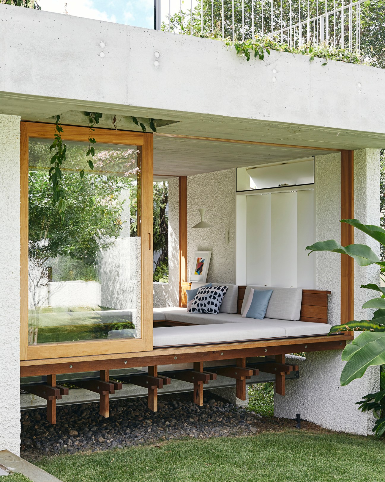 A modernist covered outdoor living space with timber framing, concrete walls, and cushioned seating overlooking a garden.