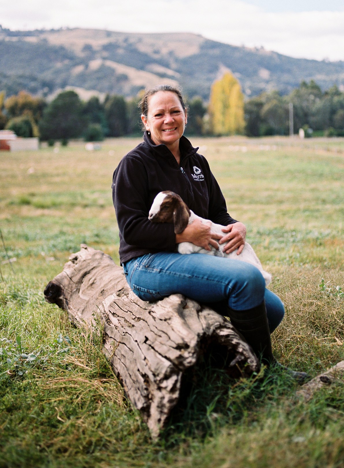 Woman in black jacket sitting on fallen log holding young goat in rural field with hills in background