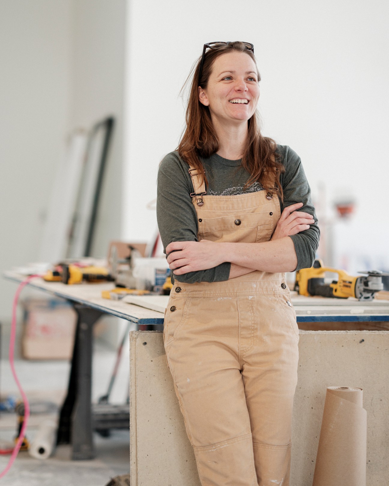 Sarah Wood standing in modern workshop space wearing overalls and green shirt with arms crossed