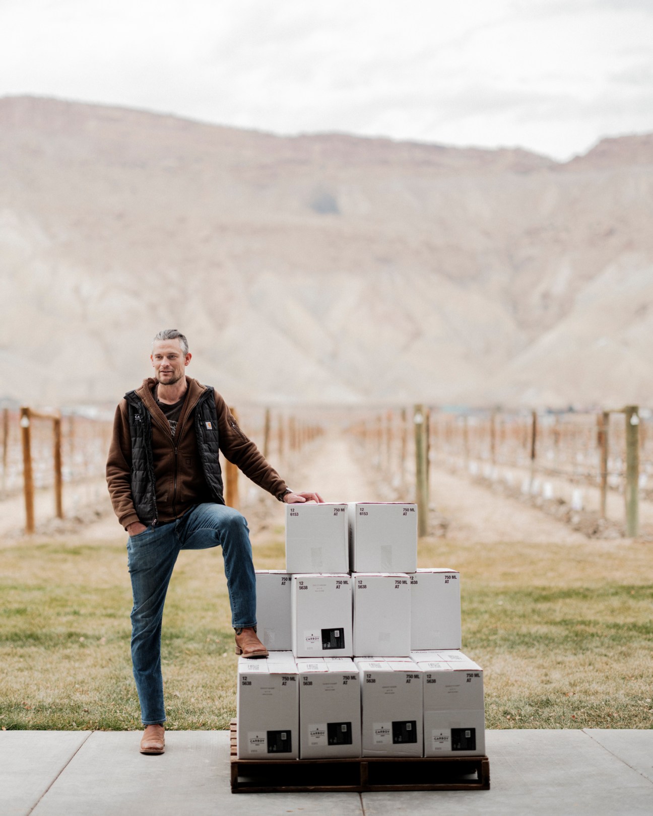 Man in brown jacket and jeans posing with stacked wine boxes in rural mountain landscape