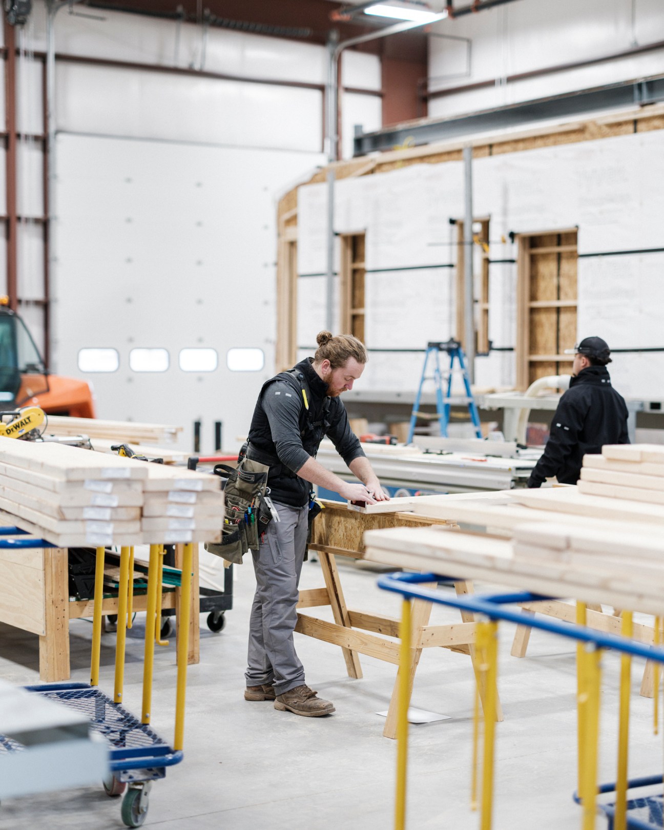 Worker in tool belt measuring wood beam in prefabricated house construction facility with exposed ceiling structure
