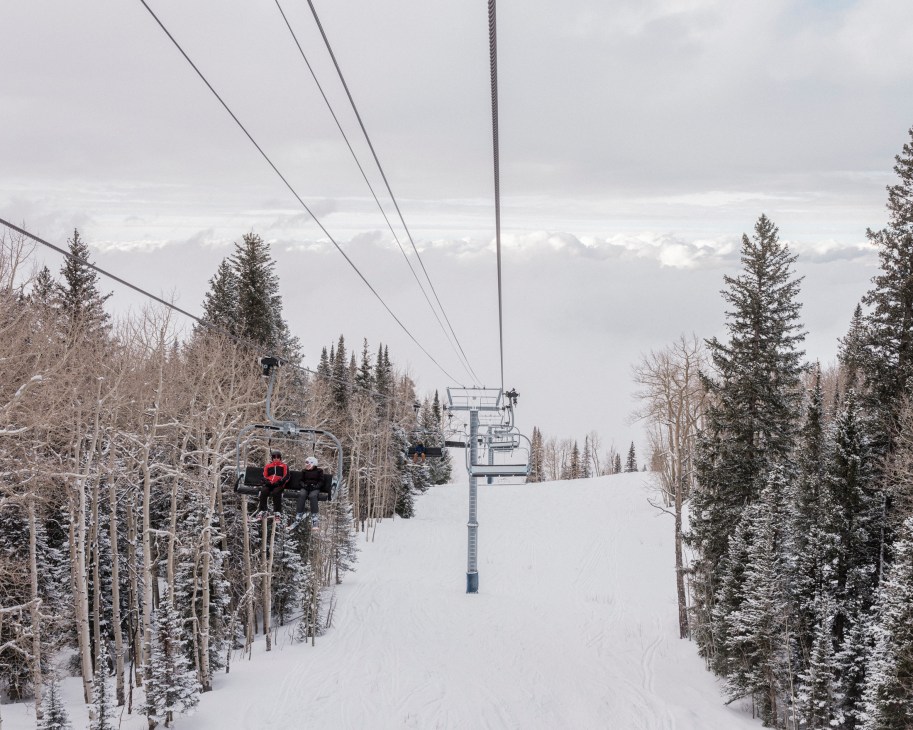 Skiers riding chairlift up snowy mountain slope at ski resort with snow-covered trees