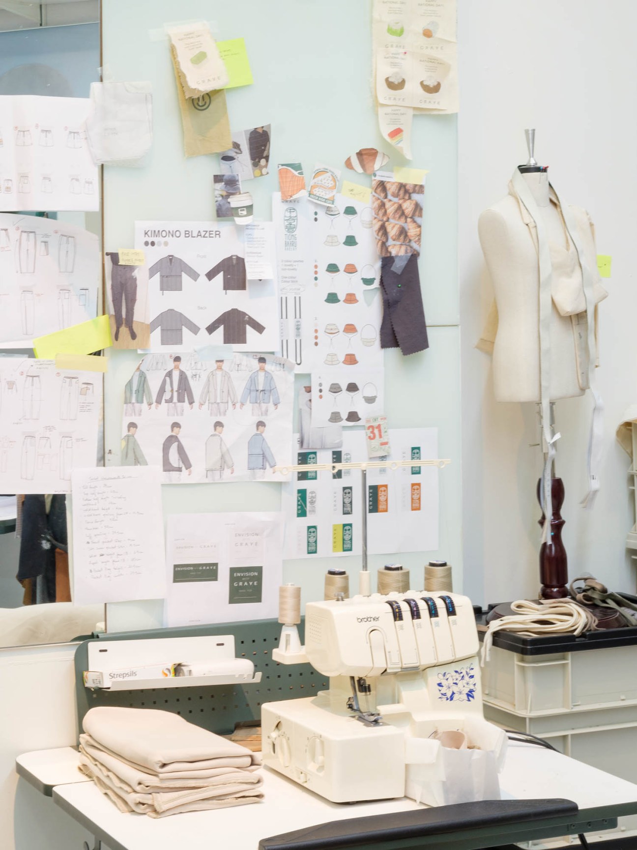 Fashion design studio workspace with sketches pinned to wall, dress forms, sewing machine and fabric samples