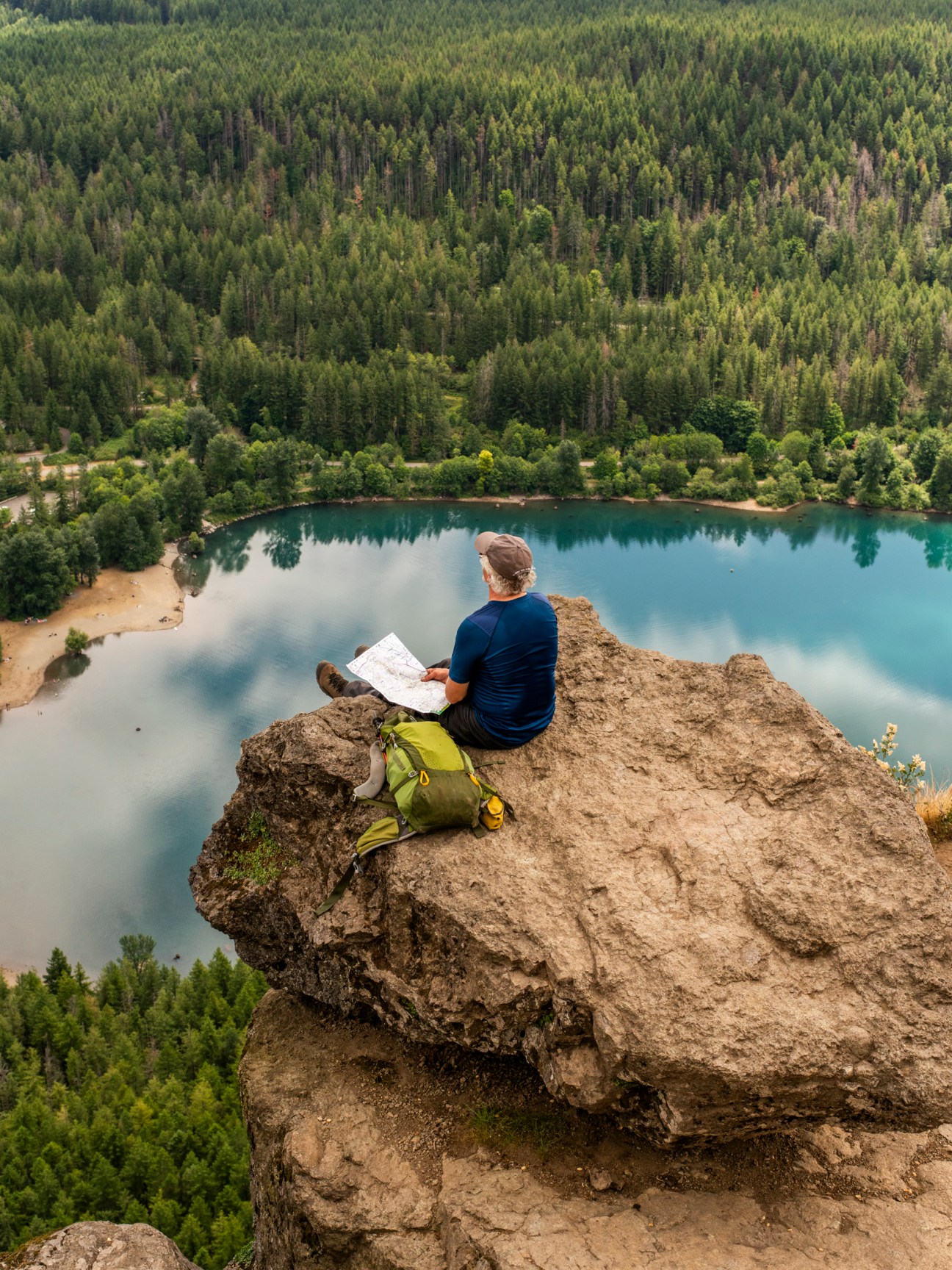 Hiker sitting on rocky outcrop consulting a paper map overlooking a turquoise lake and forested landscape.