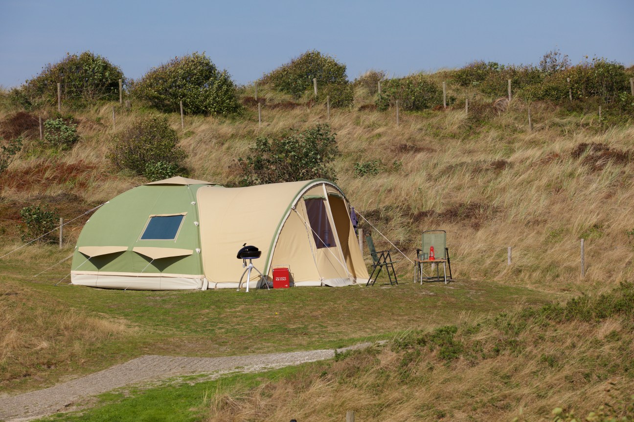 A green and cream colored tent pitched in a grassy hillside camping area with vegetation in the background.