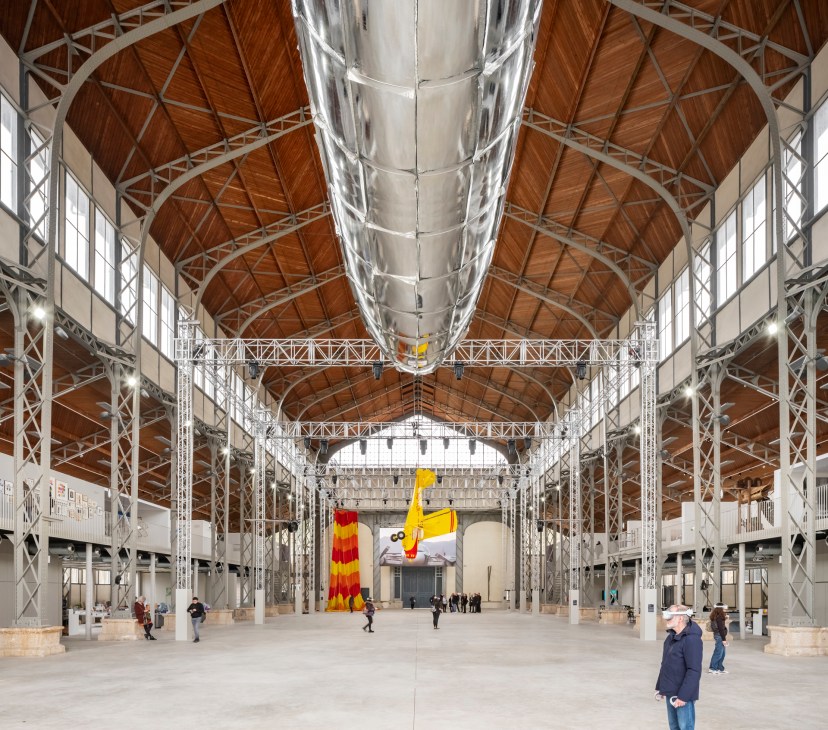 Interior of Hangar Y, a vast industrial hall with exposed steel beams and wooden roof trusses, featuring colorful fabric sculptures.