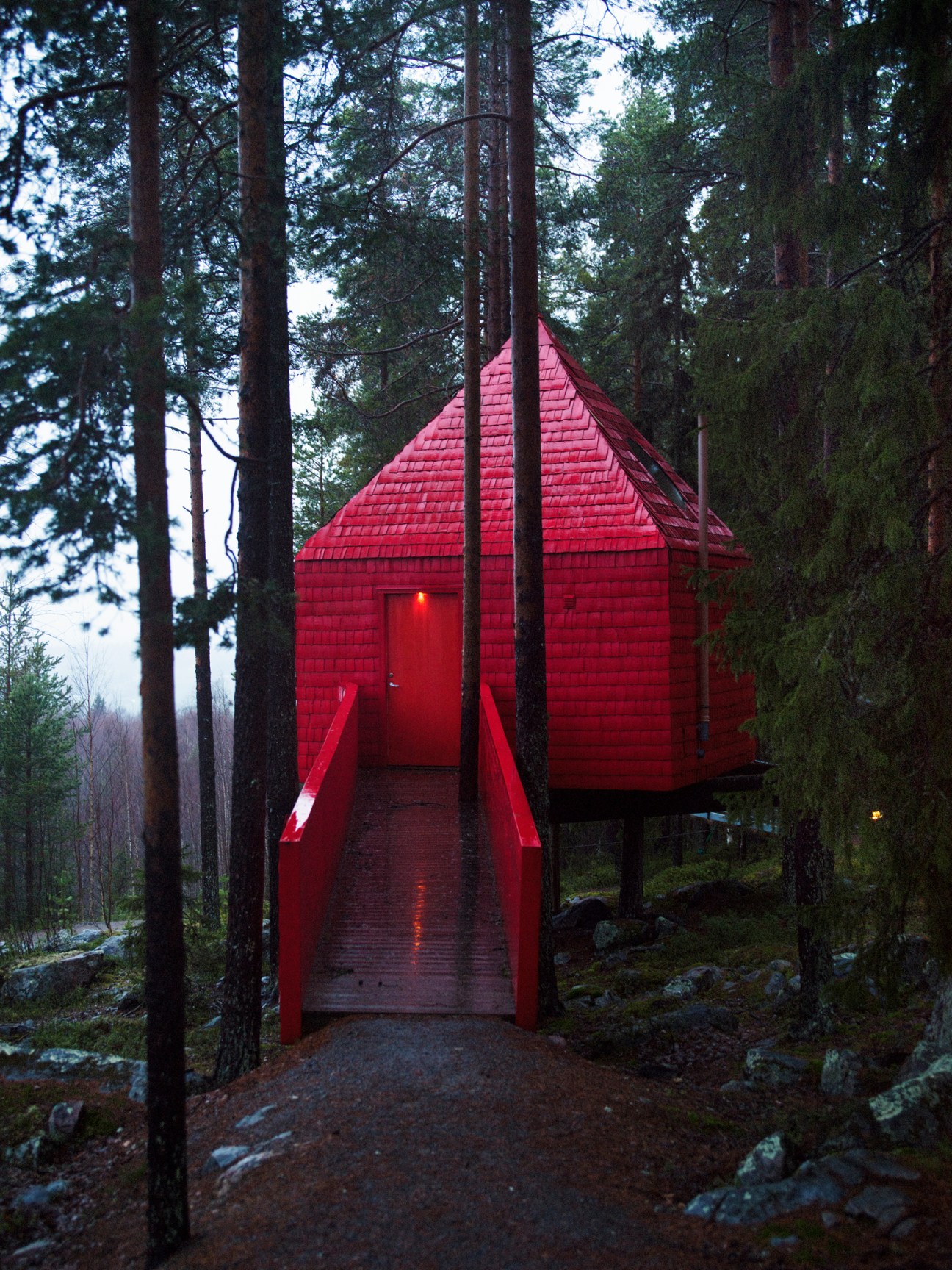 Red wooden treehouse cabin elevated on stilts in forest with illuminated entrance and walkway at dusk