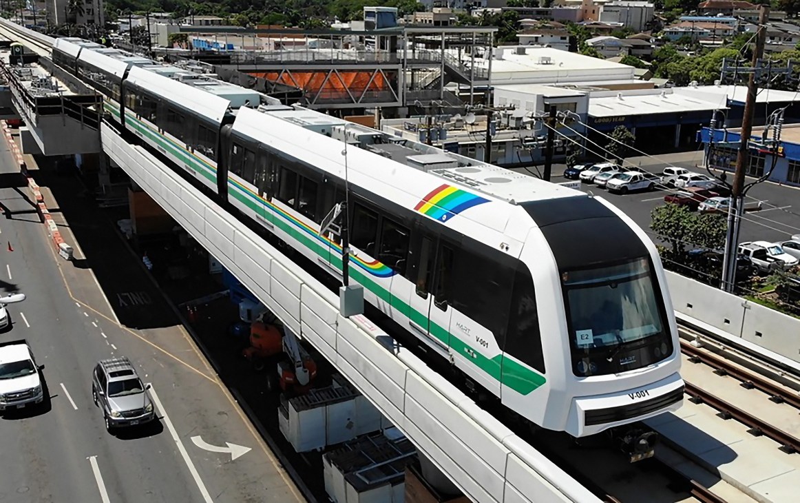 Honolulu's new white and green elevated light rail train with rainbow pride flag on side, stationed above urban street with parked cars and industrial buildings