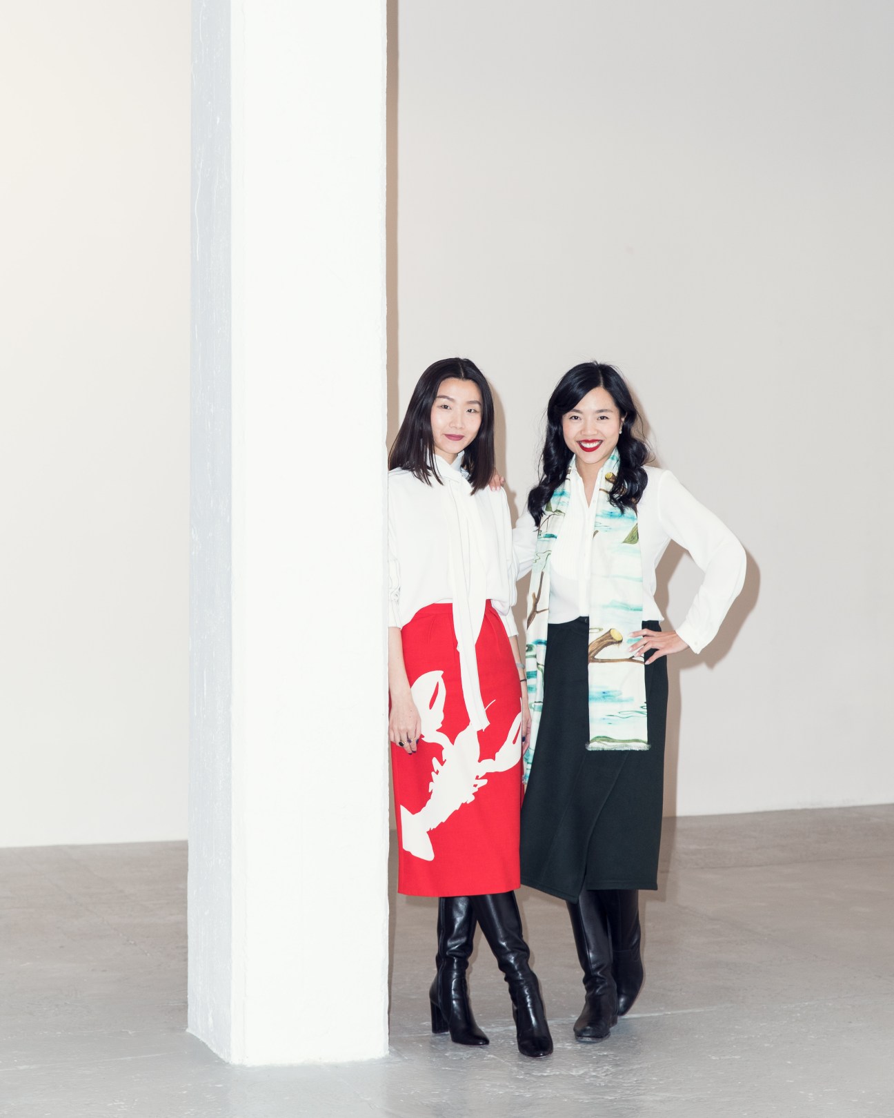 Two women standing in a gallery space next to a white column, wearing white blazers and colorful patterned skirts.