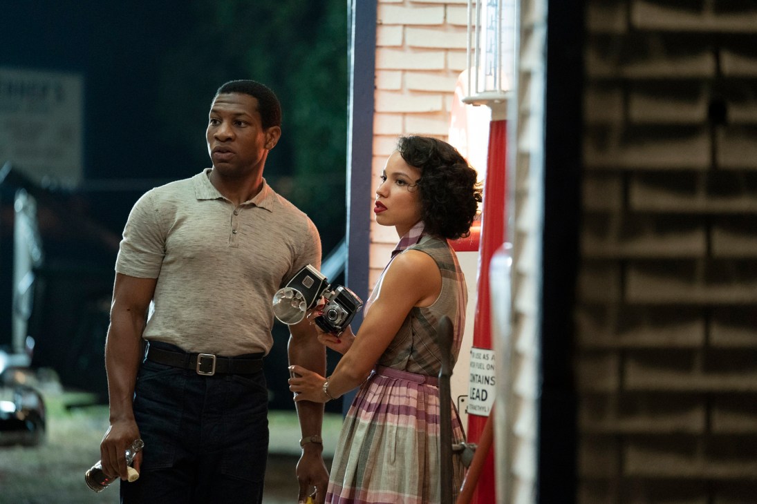 Two people in 1950s attire standing on a street, man in polo shirt and woman in plaid dress holding a camera