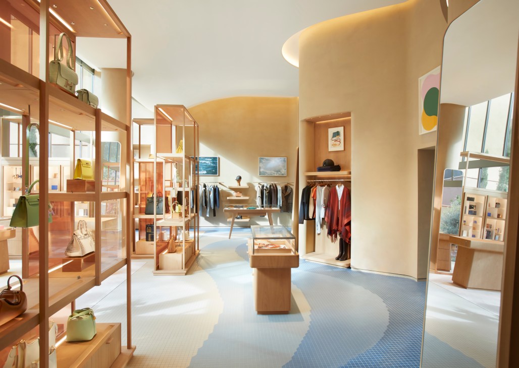 Hermès Barcelona store interior with warm sandy stucco walls, wooden display fixtures, clothing racks, and mosaic flooring with wave patterns.