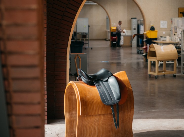Black leather horse saddle displayed on curved wooden stand in modern Hermès factory with people working in background