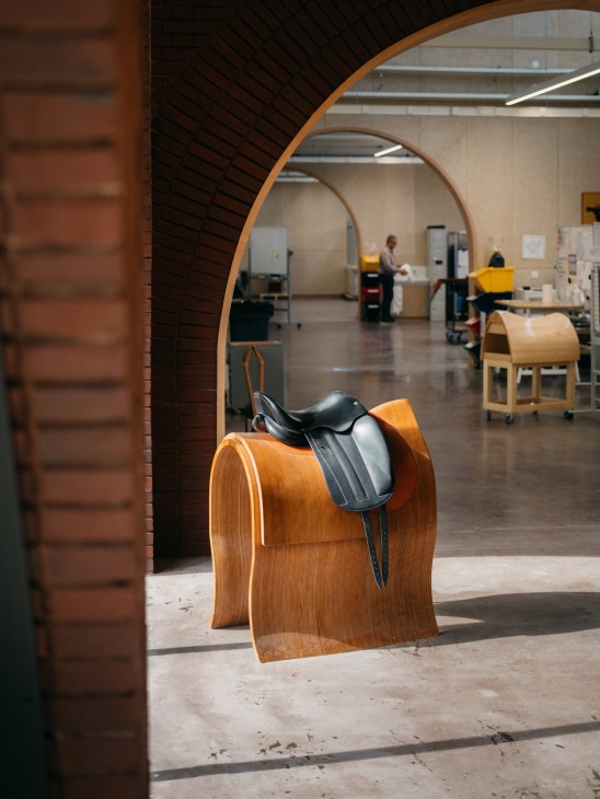 Black leather horse saddle displayed on curved wooden stand in modern Hermès workshop with brick archway