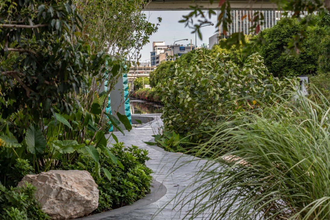 Winding wooden boardwalk through lush tropical vegetation at Chong Nonsi Canal Park with urban buildings visible