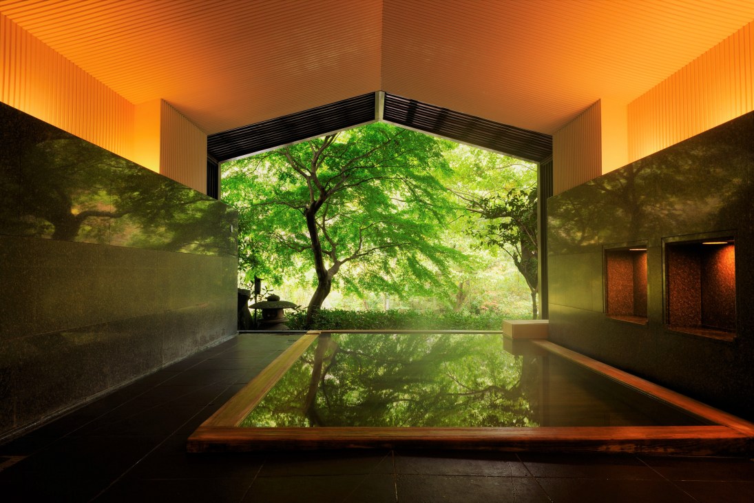 A serene indoor hot spring pool with amber-lit ceiling overlooking lush green trees through a large window frame.