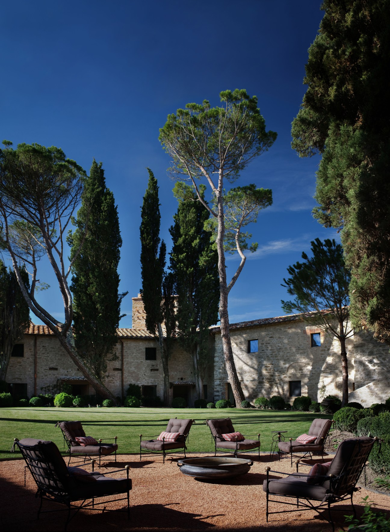 Outdoor seating area with chairs arranged in circle around fire pit with stone building and tall trees behind
