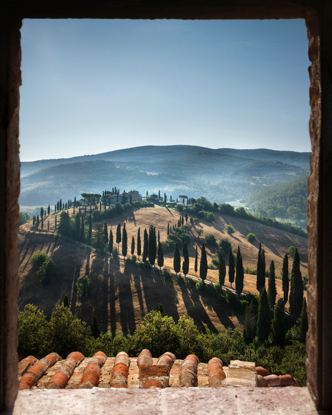View through stone window frame showing Tuscan countryside with cypress trees and rolling hills