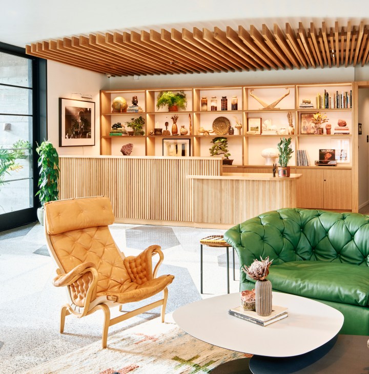 Modern hotel lobby with wood-slatted ceiling, built-in shelving, tan leather chair and green tufted sofa