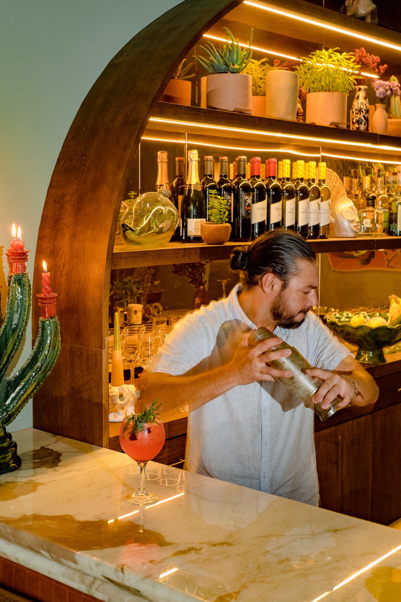 Bartender preparing cocktail at curved wooden bar with wine bottles and plants on illuminated shelves