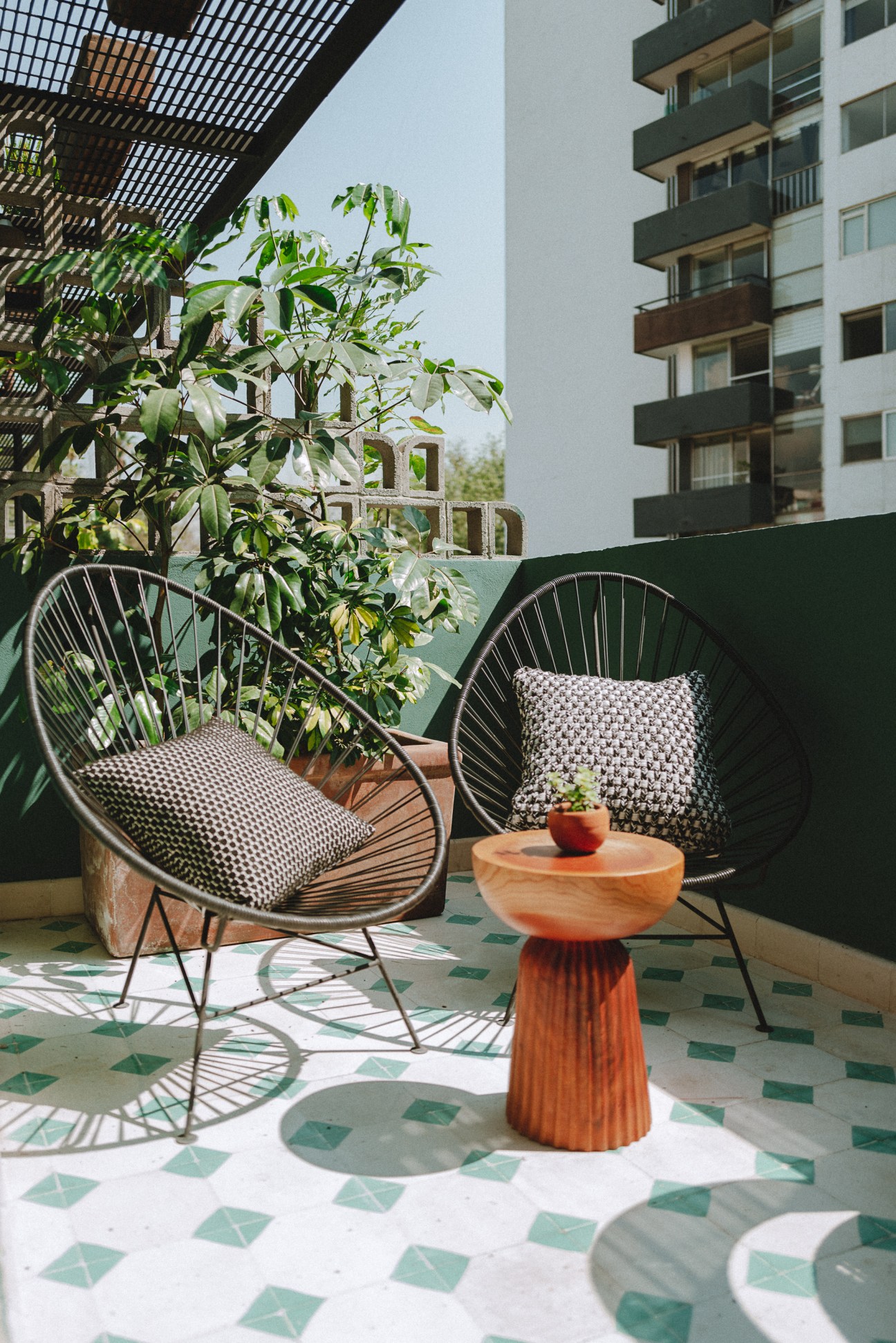 Outdoor terrace with two black wire chairs, wooden side table, and green geometric tile floor overlooking city buildings